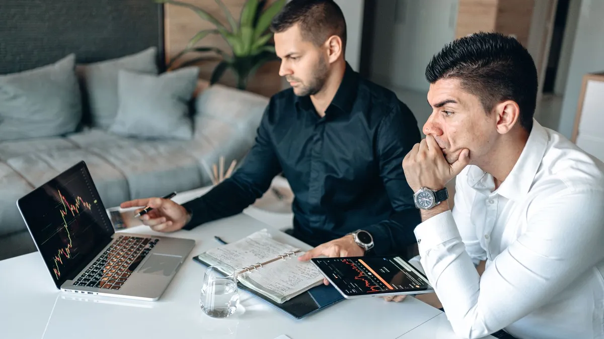 Two men reviewing a home budget with a laptop, tablet, and notebook